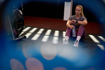 Sporty woman sitting on the gym floor, checking her smartwatch after a workout, with a water bottle...
