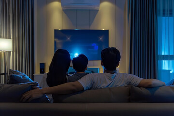 Rear view of father, mother, and son are sitting on sofa while watching television at night