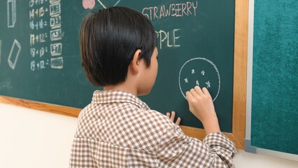 Asian boy drawing picture to express and develop imagination while standing at blackboard in classroom. Attractive elementary student writing at blackboard in art lesson. Education concept. Pedagogy.