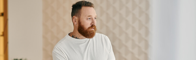 A man with a red beard lost in thought, in a modern living room setting.