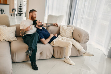 Obraz premium Two men in casual clothes, a happy gay couple, sit on a modern couch in a living room, sharing a moment of love and connection.