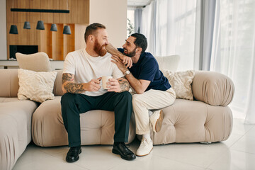 A happy gay couple in casual clothing sit closely together on a couch in a modern living room,...