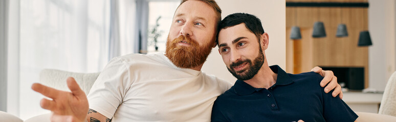 Two bearded men in casual attire sit closely on a couch in a modern living room, enjoying quality...