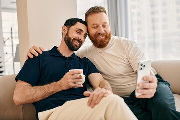 Two happy gay men in casual clothes sit on a couch, engrossed in their cell phone, sharing a moment...