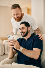 A happy gay couple in casual clothes sits on a couch, engrossed in a cell phone, enjoying a moment of modern connection.