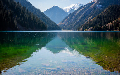 a serene alpine lake with crystal-clear waters reflecting the sky, bordered by dense coniferous...