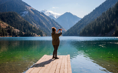 a adult woman standing on a wooden dock at the edge of an alpine lake, taking a photo with their...