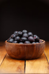 A wooden bowl filled with 'Indian blackberry' or Jamun (Syzygium cumini) fresh fruits, on a dark background. Front view.