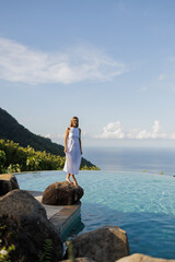 Woman in  white dress walking on the edge of an infinity pool,  sea and palms on background. Summer...