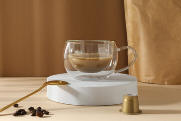 Golden coffee capsule, spoon and glass cup with coffee drink on beige background