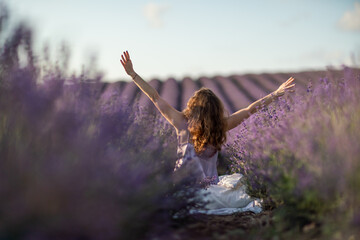 A woman is sitting in a field of lavender flowers. She is stretching her arms out in front of her.