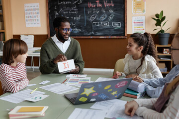 Young African American teacher of English sitting at table in classroom showing alphabet cards to kids during lesson © AnnaStills