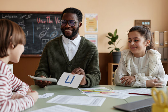 Cheerful African American male teacher of English working with group of kids showing phonics cards during lesson in classroom