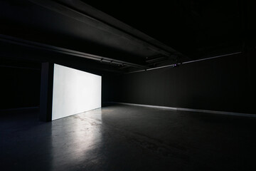 Big blank white illuminated screen backdrop in large empty dark room concrete floor. Nobody in room.