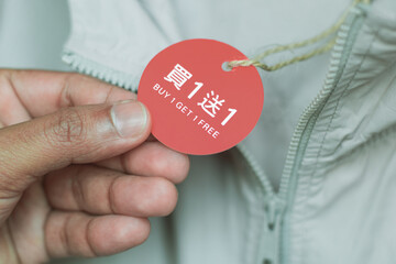 Close-up of a man hand holding a hang tag of a jacket displaying buy 1 get 1 free offer both in...