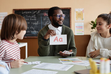 Medium shot of cheerful African American teacher of foreign language sitting at table in classroom...