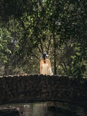 A young woman girl in a dress and hat from behind back standing on a rock stone bridge in a forest...