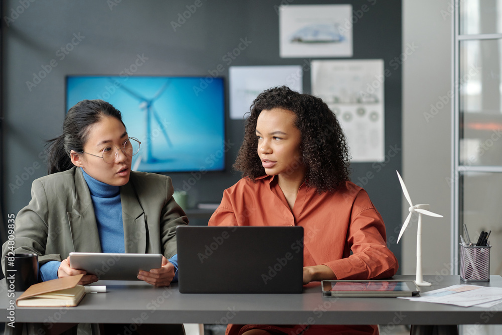 Poster two young successful intercultural female employees looking at one another while discussing online d