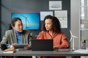 Two young successful intercultural female employees looking at one another while discussing online data and preparing new project