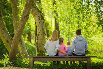 Mother, father, daughter and little son sitting on bench and staring at green leaves, grass and trees at beautiful park in sunny spring day. Family spending time together in nature. Back view.