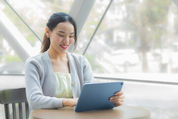 Fototapeta premium Asian professional business female in light gray long sleeve shirt is looking at tablet or notepad confidently in her hands while she is sitting working at coffee shop among business background city.