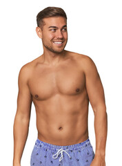 Fototapeta premium Young man shirtless in swimwear, studio looks aside smiling, cheerful and pleasant.