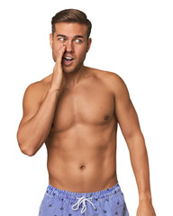 Fototapeta premium Young man shirtless in swimwear, studio is saying a secret hot braking news and looking aside