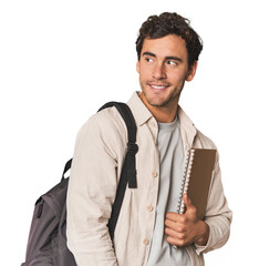 Young Hispanic student with notebook looks aside smiling, cheerful and pleasant.