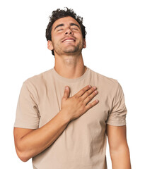 Young Hispanic man in studio laughs out loudly keeping hand on chest.