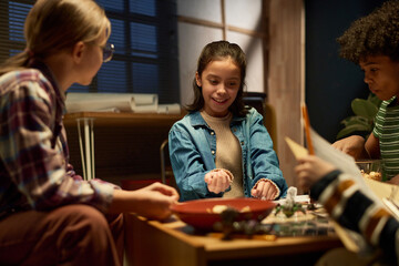 Smiling pretty girl with dice in hand sitting by table in front of her friends and looking at board while playing tabletop game
