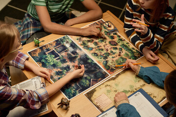 High angle of four intercultural youngsters gathered by table with new board game and playing while...