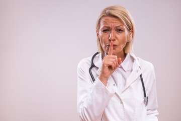 Portrait of mature female doctor showing silence sign on gray background.