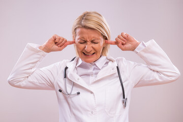 Portrait of  overworked mature female doctor with hands in ears shouting on gray background.