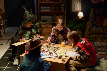 Four intercultural youthful children in costumes of board game characters sitting by table and...
