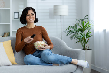 Relaxed and smiling young woman sitting and relaxing on the sofa at home, holding a plate with...