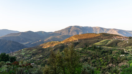 tranquil landscape at golden hour, with rolling hills and mountains bathed in soft sunlight. A tapestry of greenery and cultivated fields is interspersed with winding roads and sparse buildings.