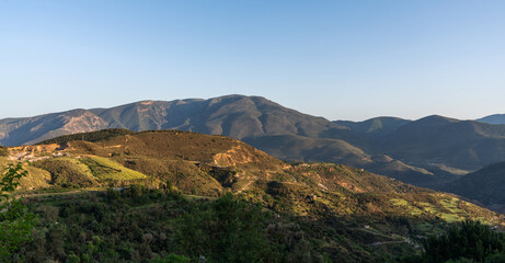 a vast landscape with layered mountain ranges, the sunlight casting soft shadows and highlights on the ridges, with lush greenery in the foreground under a clear sky.