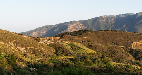 a vast landscape with layered mountain ranges, the sunlight casting soft shadows and highlights on the ridges, with lush greenery in the foreground under a clear sky.