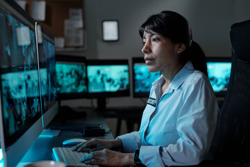 Young serious female in security uniform sitting in armchair in front of computer in surveillance...