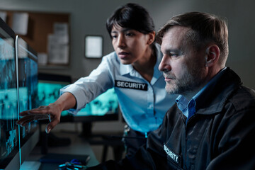 Serious mature security guard looking at computer screen while his colleague pointing at it during...