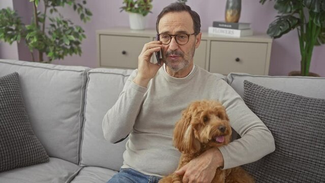 Mature man talks on phone while cuddling labradoodle on couch indoors