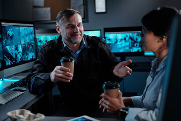 Smiling mature male security guard with cup of coffee looking at young female colleague during...