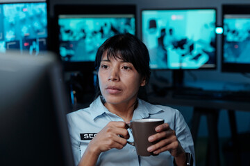 Young Hispanic female officer working in surveillance room holding cup of coffee and watching cctv...