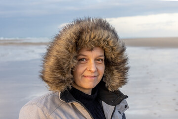 A woman in a fur hooded jacket smiles warmly on a cold beach day, under a cloudy sky and by the...