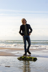 A relaxed man in jeans and jacket stands on a rock by the beach, with waves in the background under...