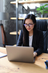 A young Asian entrepreneur, focused and confident, utilizes her laptop in a modern office setting.