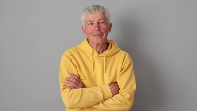 Approved gray haired Caucasian mature man wearing yellow hoodie isolated over gray background standing with crossed arms nodding yes and agree gesture