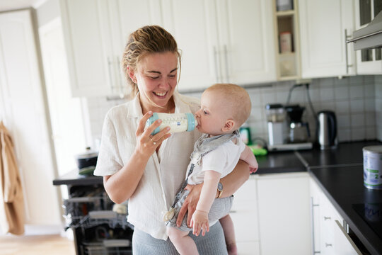 Smiling young woman feeding toddler son with milk bottle in kitchen at home