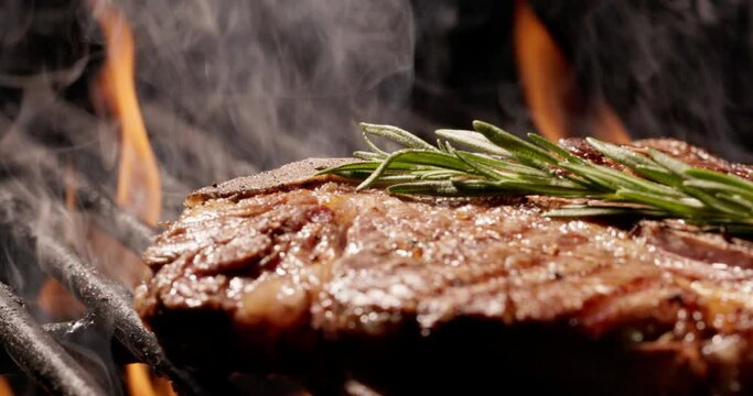 Close-up. Slow motion. T-bone steak on grill, juicy and appetizing, covered with crispy golden crust, seasoned with sprig of rosemary to reveal flavor. Slow flames on dark background, cinematic AD