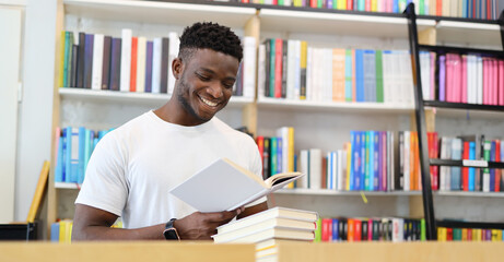 Cheerful student studying in library, holding book, sitting by bookshelf, smiling, focused on...